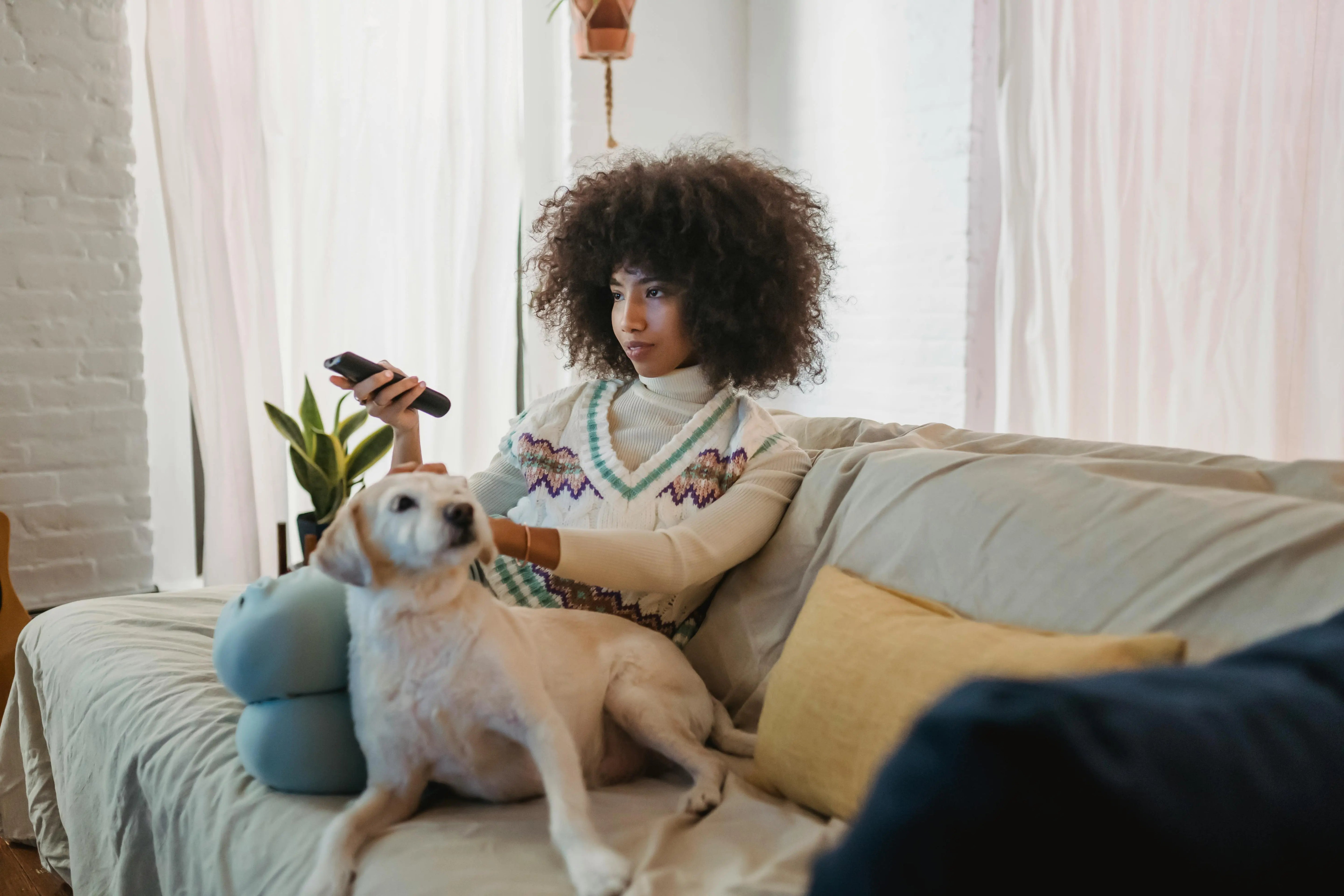Woman sitting on a couch with a dog, holding a remote control, in a cozy living room.