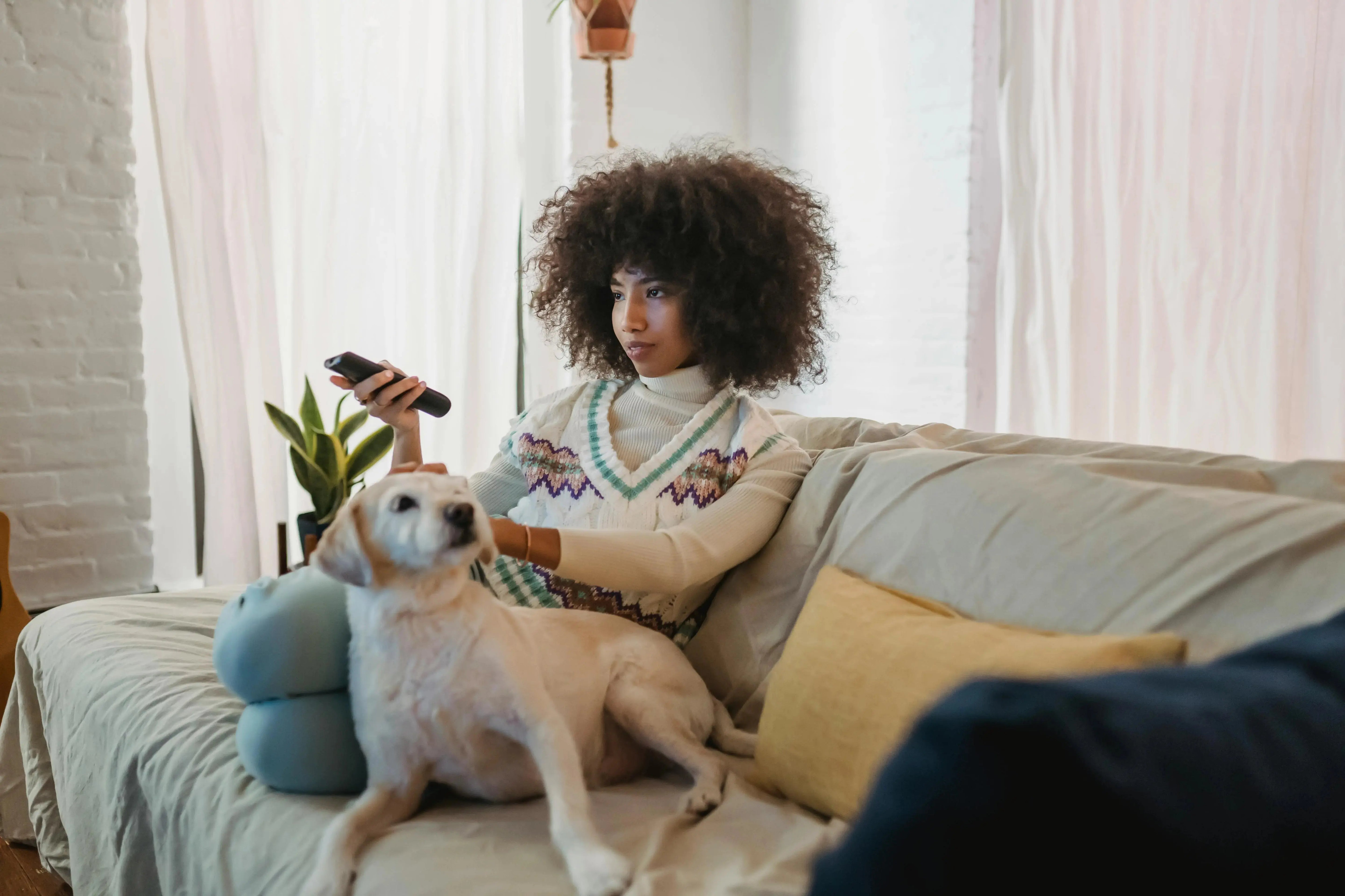 Woman sitting on a couch with a dog, holding a remote control, in a cozy living room.